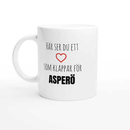 Vit kaffemugg med texten 'Här ser du ett hjärta som klappar för Asperö' och ett rött hjärta. En stilren kopp för alla som älskar Asperö i Göteborgs skärgård.