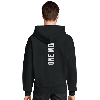Manlig modell som visar det stora vita ryggtrycket "ONE MORE" på en svart oversized hoodie. Modern passform för träning.