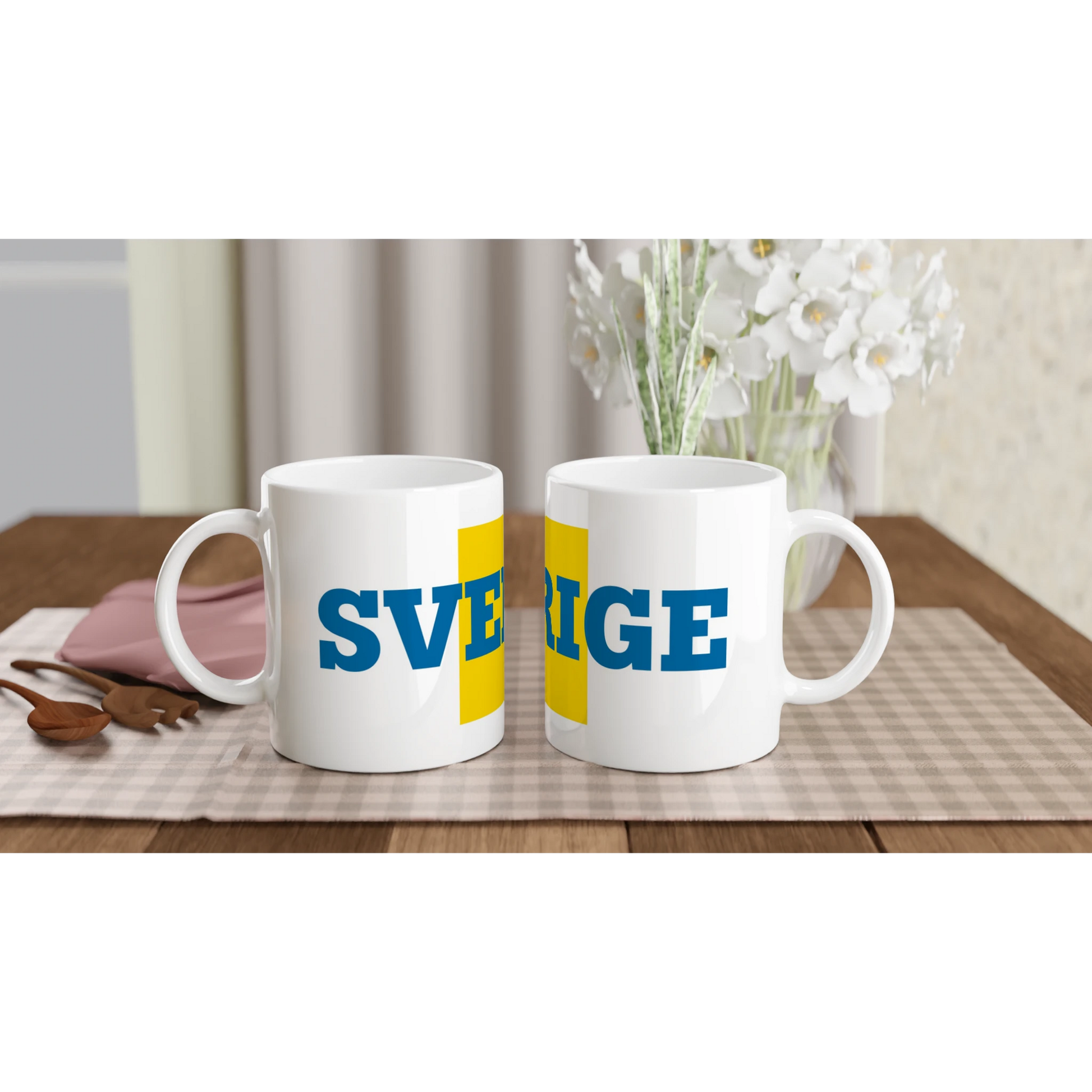 Två Sverige-muggar uppställda på ett bord för en svensk fika. Visar muggarnas design i en hemtrevlig miljö.